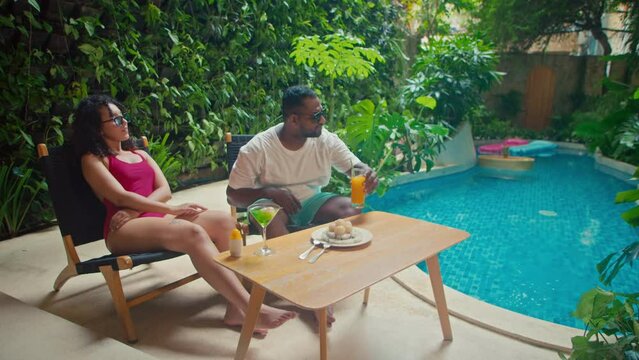 Full shot of young multiethnic couple in swimming suits and sunglasses chilling out and sunbathing next to pool at luxury holiday villa, talking and drinking cocktail and fruit juice