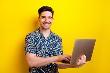 Portrait of handsome satisfied guy with trendy hairstyle wear print shirt hold laptop chatting isolated on yellow color background