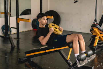 A young handsome asian man does a set of weighted situps with a yellow barbell plate on a decline...
