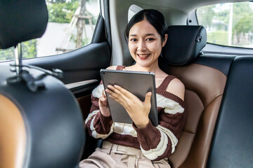 Happy young asian woman passenger sitting in a car plays with a digital tablet, typing on tablet, sits in the back seat of a car.