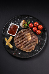 BBQ beef steak with sauce on a black plate with vegetables, tomatoes and corn. Black background. Restaurant supply.