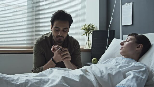 Medium shot of Middle Eastern young man sitting in hospital ward holding hand of his Caucasian girlfriend lying in coma in bedMedium shot of Middle Eastern young man sitting in hospital ward holding h