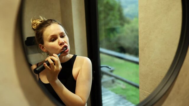 the girl brushes her teeth with an electronic black brush in the bathroom. woman looks in the mirror in the morning. in the background the forest in the window.