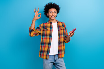 Photo of satisfied guy with perming coiffure dressed plaid shirt holding smartphone show okey approve isolated on blue color background