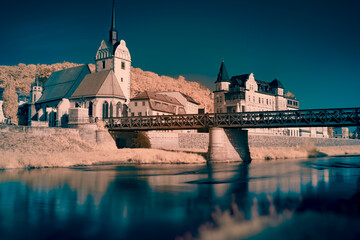 An infrared photo of a bridge over a river with a castle in the background. The bridge is arched...