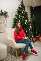 Beautiful smiling young girl in red sweater sitting in chair near Christmas tree.  Happy New Year. Fashion woman relaxing in warm and cozy evening at home. Decorated interior living room of house.