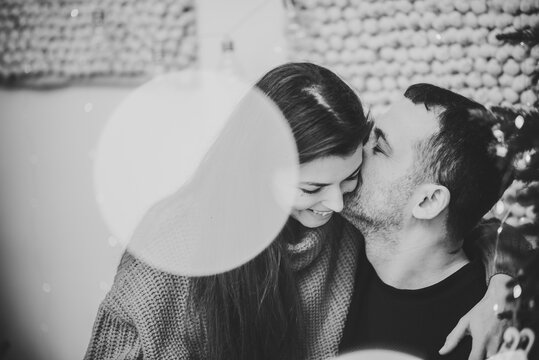 Man Kiss Woman Near Christmas Tree Decorated Garlands In Interior Room. Couple Enjoying Winter Time Together On Holiday. Happy New Year. Xmas Morning At Home. Closeup Love Bokeh. Black And White Photo