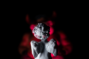 A toy white angel with wings against the background of a huge red demon with horns. The angel sits...