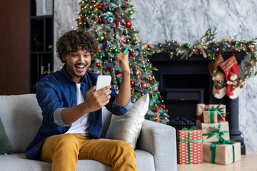 Successful man received online notification of victory win, winner for christmas sitting near tree living room on sofa in house indoors, hispanic using app on smartphone, holding hand up achievement.