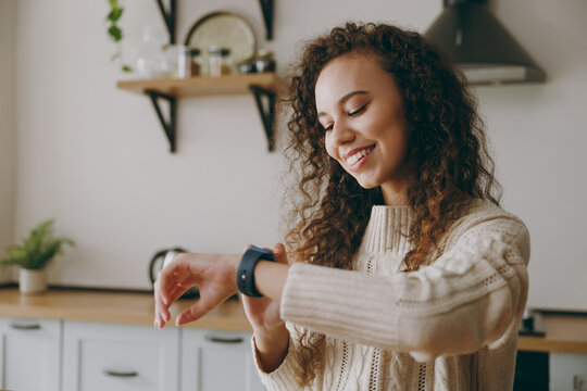 Young smiling happy housewife woman of African American ethnicity wear casual clothes sweater loot at smart watch check time sit at table in light kitchen at home alone Lifestyle cooking food concept