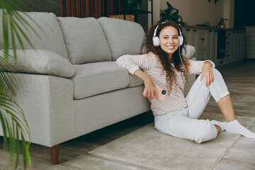 Full body young woman in casual clothes headphones sit near grey sofa use mobile cell phone listen music stay at home hotel flat rest relax spend free spare time in living room indoor Lounge concept