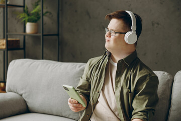Young man with down syndrome wear clothes headphones listen music use mobile cell phone sit on grey...