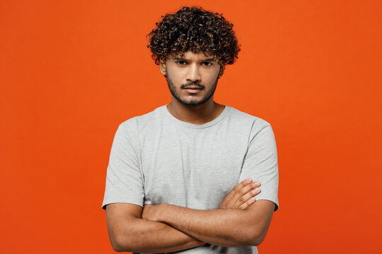 Young sad frowning mad furious shrewd Indian man he wearing t-shirt casual clothes hold hands crossed folded looking camera isolated on orange red color background studio portrait. Lifestyle concept.