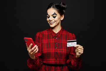 Young woman with Halloween makeup face art mask in clown costume red dress hold use mobile cell phone credit card isolated on plain solid black background studio portrait Scary holiday party concept