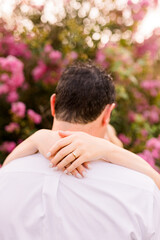 Couple are embracing, her hand is over his neck, showing off her engagement ring.