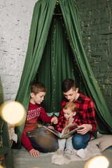 Obraz na płótnie Canvas Childs enjoying time together. Merry Christmas. Children reading books for little brother on floor. Large big family with three kids. Interior with canopy with garlands. Concept of winter holiday.