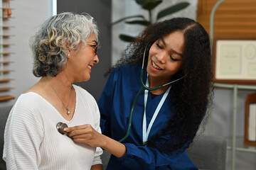 An African-Asian doctor wearing a scrub uniform listening to an older woman lung or heart sounds with a stethoscope during a visit at home, medical checkup and health care concept