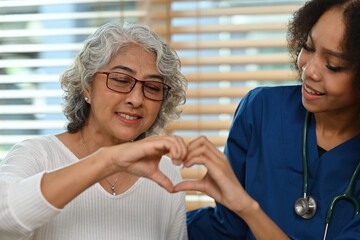 Portrait image of an old Asian woman and African nurse or caregiver making a heart shape with hands together, Care and health insurance for the elderly concept