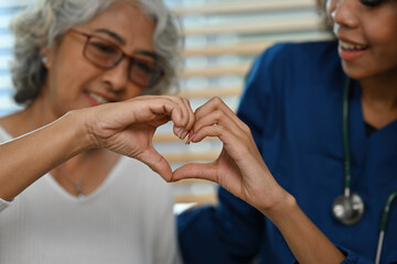 Close-up shot image of an old Asian woman and African nurse or caregiver making a heart shape with hands together, Care and health insurance for the elderly concept.
