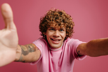 Handsome young curly hair man stretching out hands and smiling while making selfie against pink background
