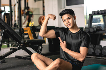 Fototapeta na wymiar A young handsome and confident asian man flexing his bicep while sitting on the mat at the gym.