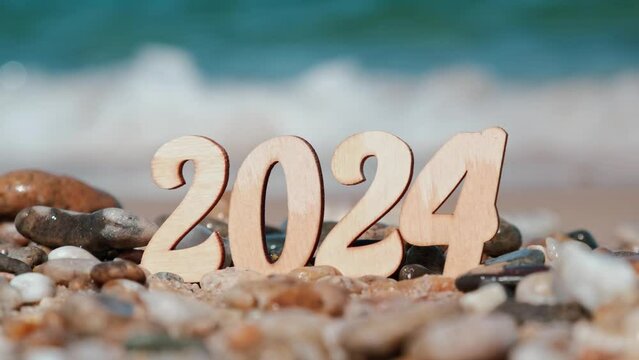 closeup of wooden New Year numbers 2024 on beach by sea against background of waves and surf. big wave covers and washes away numbers. concept of tourism, relaxation and travel. plans for future.