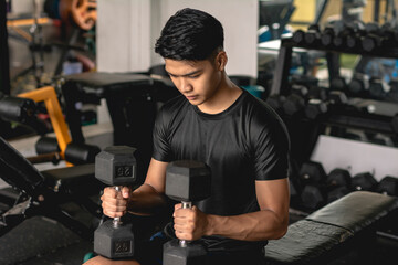 A young asian man looking focused placing two dumbbells on his knees while preparing to do a set of...