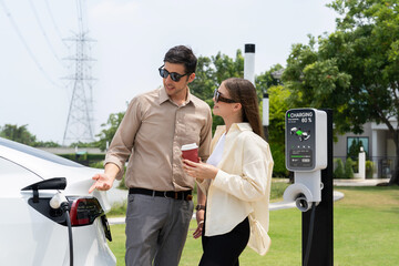 Young couple with coffee pay for electricity while recharge EV car battery at charging station connected to electrical power grid tower as electric industrial for eco friendly car travel. Expedient