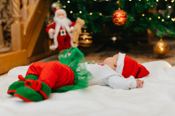 A newborn baby in a Christmas elf costume is lying on a back on a white blanket and looking at a Christmas tree with bokeh for Christmas. No face. 