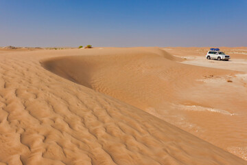 Curved dune with undulating red sand in the foreground, white car with luggage in the Rub Al Khali desert in the background. Oman.