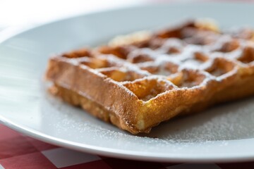 A close up portrait of a belgian tasty delicious waffle with powder sugar as a topping on it, lying on a white plate on a red table cloth ready to be eaten during breakfast or lunch or as a snack.