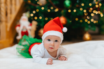 Fototapeta na wymiar Baby girl with Christmas elf clothes is lying on a white blanket near Christmas tree with bokeh. Christmas concept. Infant baby in her first Christmas. Funny baby. 