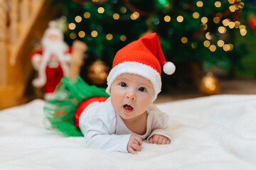 Baby girl with Christmas elf clothes is lying on a white blanket near Christmas tree with bokeh. ...