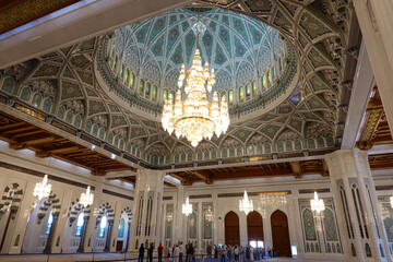 Obraz premium Prayer hall with white marble side colonnade and ornate dome on the ceiling and chandelier in the centre, in the background tourists queuing up at the great mosque of Sultan Qaboos. Oman.