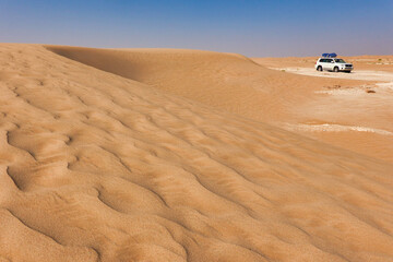 Curved dune with undulating red sand in the foreground, white car with luggage in the desert in the background.