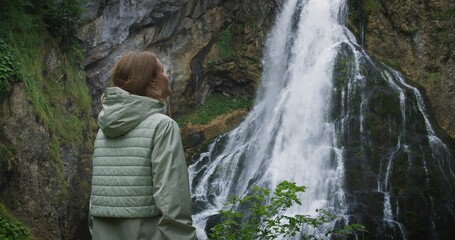 Woman enjoying a mighty waterfall at rainy day. Girl traveler in autumn coniferous forest....