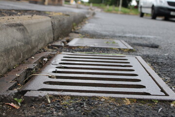 Grate of a storm sewer drain up close perspective, blurred street in background - obrazy, fototapety, plakaty