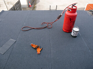 Red propane gas cylinder with connected blowtorch standing on almost finished new bitumen roofing...