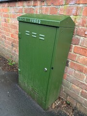 Green fibre communications cabinet installed on a public pavement, against a brick wall, taken on a cloudy afternoon