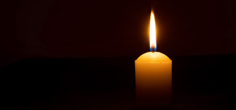 Single burning candle flame or light glowing on a big white candle on black or dark background on table in church for Christmas, funeral or memorial service with copy space.