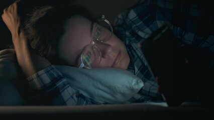 Device. A young girl in night pajamas is upset using her phone while lying in bed in bed. Steady camera movement news watching woman looking at her phone at night. Insomnia man looking at phone