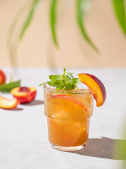 Peach tea with ice and mint. Homemade cold healthy vegetarian drink on a light background with fresh fruits, palm leaf and shadows.