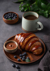 Freshly baked chocolate croissant with blueberries on a wooden plate on a dark background with a cup of black coffee close up. The concept of a delicious homemade breakfast