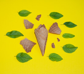 A broken ice cream cone surrounded by leaves on a yellow background. Flat lay. Summer concept.