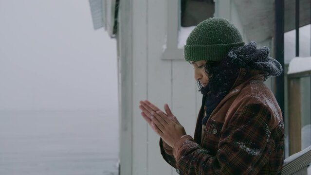 Medium profile shot of thoughtful Indian woman in plaid jacket, green hat standing near house at lake during blizzard, robbing hands, getting warm. High quality 4k footage