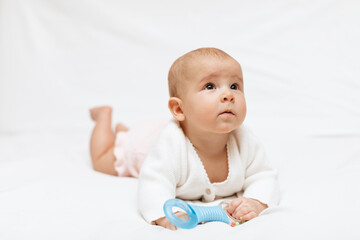 Infant baby girl in a white knitted blouse and pink knitted underpants is lying on stomach on a white blanket and playing with teething toy. Close up. 