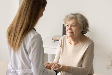 Older woman visit to doctor for medical checkup in hospital. Caring young therapist in white uniform holding hands of patient inform good news about treatment, support encouraging old person. Medicare