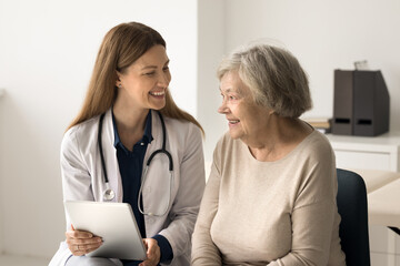 Smiling young female doctor in uniform hold digital tablet shows result of medical screening examination, give consultation, explain electronic prescription. Older patient receiving professional help