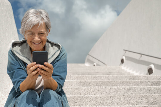 Handsome happy senior woman sitting outdoors on staircases using mobile phone apps, elderly gray haired lady enjoying tech and social