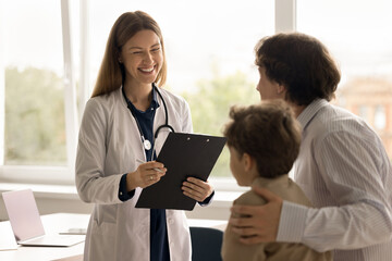 Smiling female pediatrician in uniform hold clipboard talk to little 8s patient during consultation at private clinic. Boy with dad receive professional help at meeting with doctor. Medical healthcare - obrazy, fototapety, plakaty